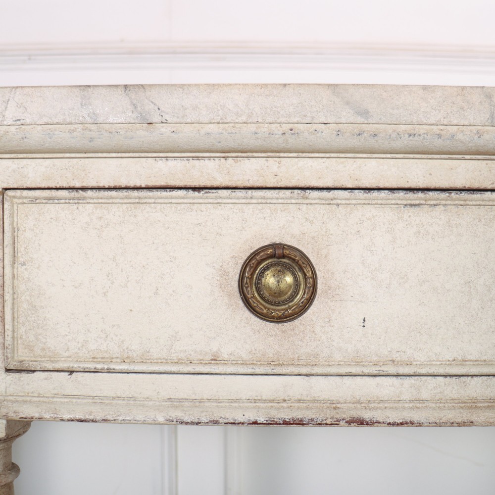 An antique off-white painted console table featuring a light marble top, three drawers with brass handles, four column supports, and a curved lower shelf. View VII