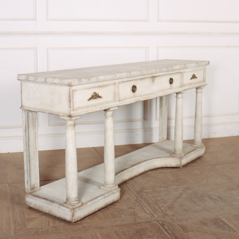 An antique off-white painted console table featuring a light marble top, three drawers with brass handles, four column supports, and a curved lower shelf. View VIII