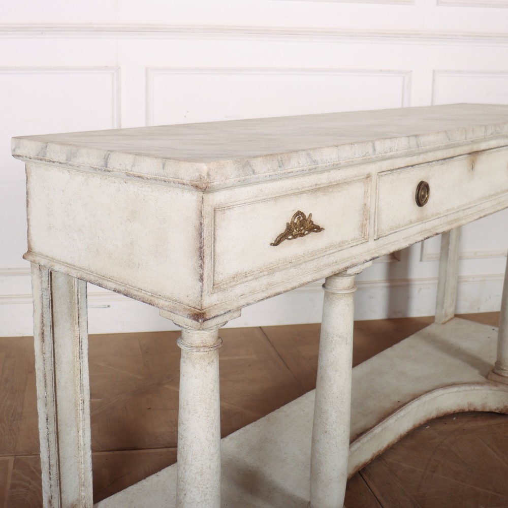 An antique off-white painted console table featuring a light marble top, three drawers with brass handles, four column supports, and a curved lower shelf. View IX