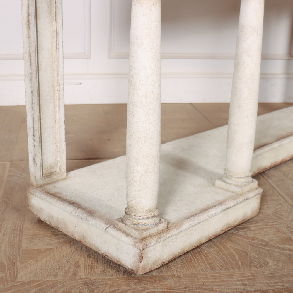 An antique off-white painted console table featuring a light marble top, three drawers with brass handles, four column supports, and a curved lower shelf. View X
