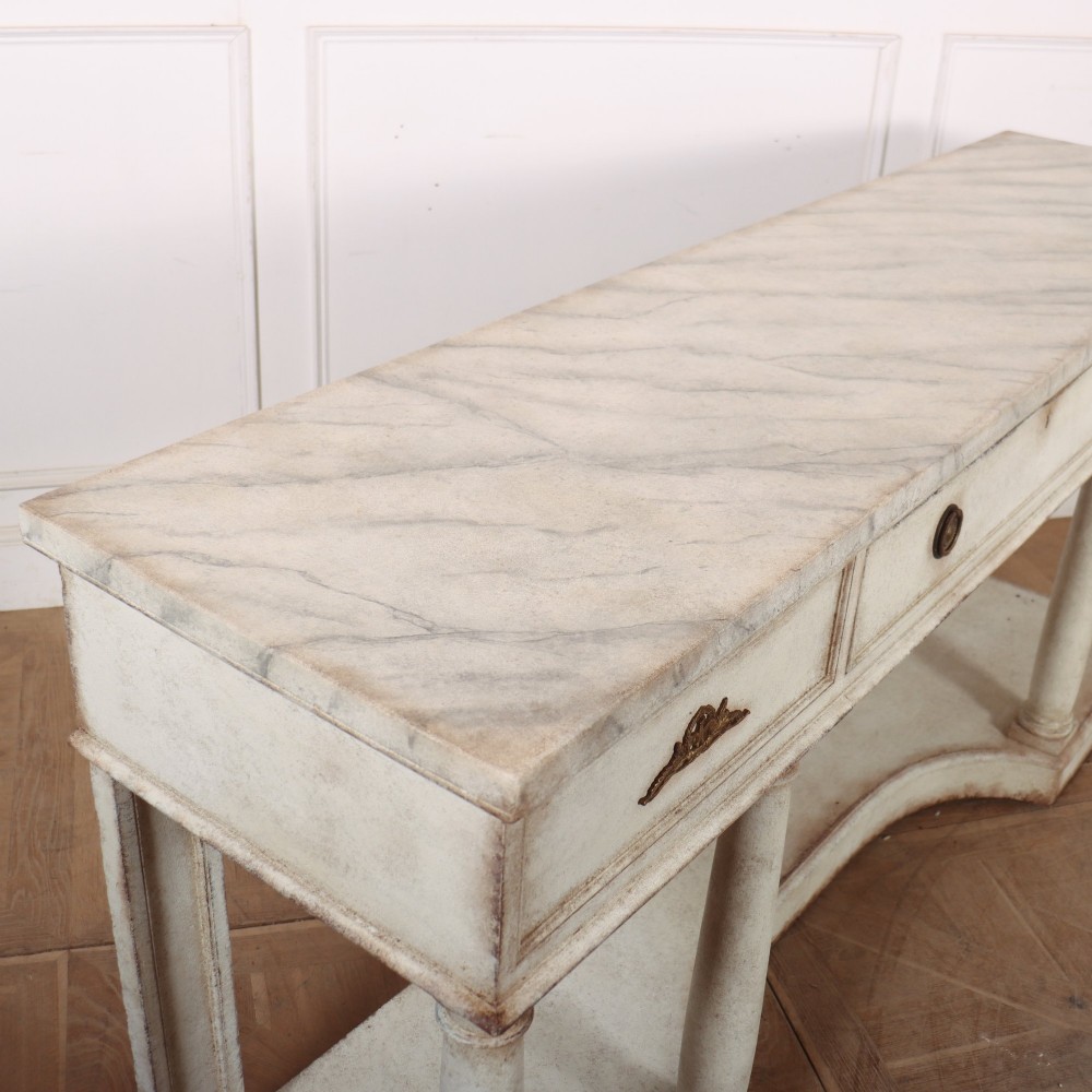 An antique off-white painted console table featuring a light marble top, three drawers with brass handles, four column supports, and a curved lower shelf. View XI