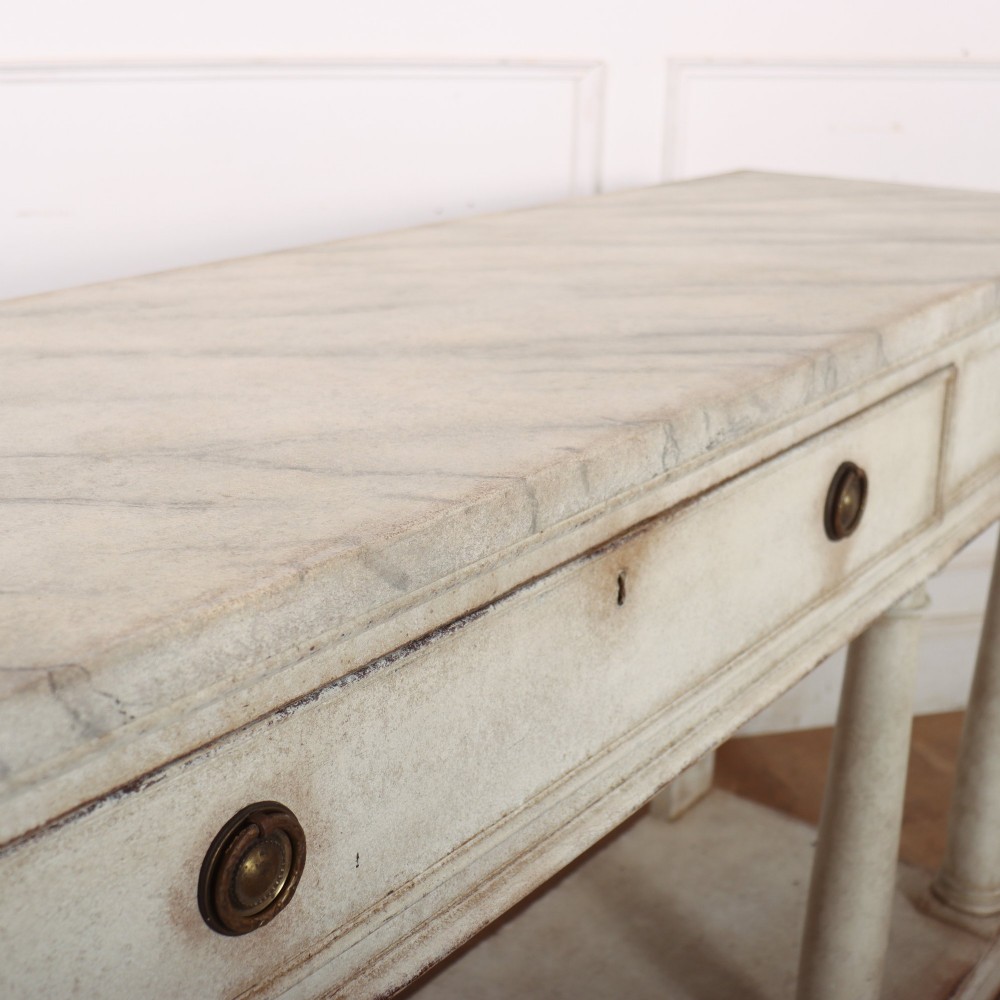 An antique off-white painted console table featuring a light marble top, three drawers with brass handles, four column supports, and a curved lower shelf. View XII