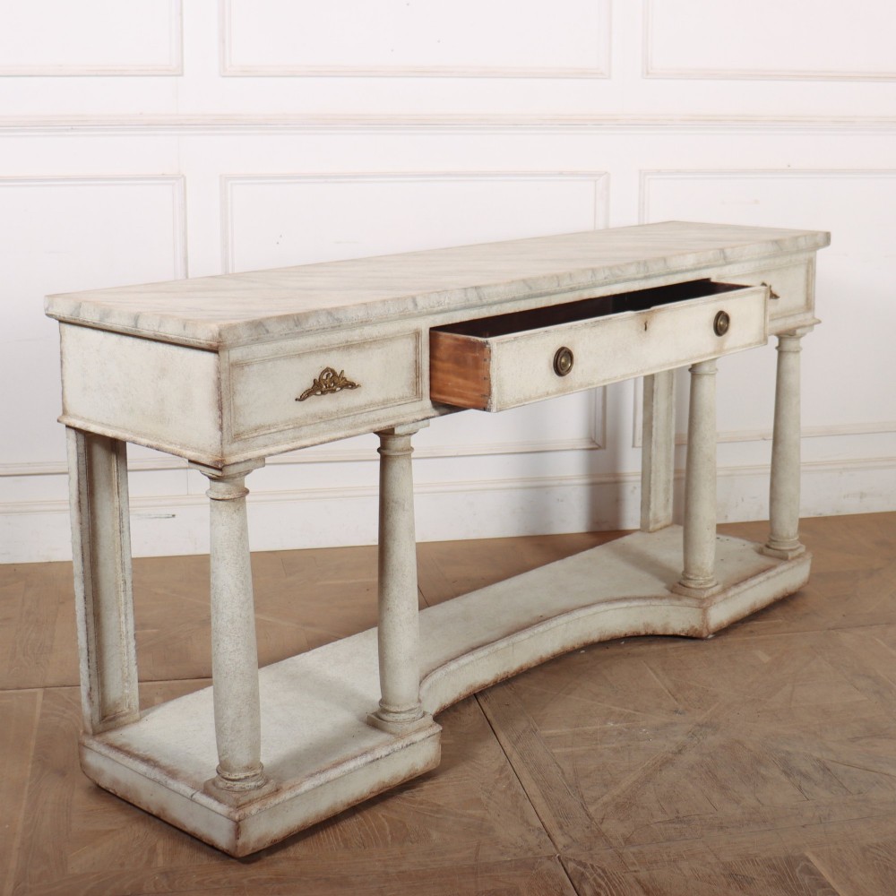 An antique off-white painted console table featuring a light marble top, three drawers with brass handles, four column supports, and a curved lower shelf. View XIII