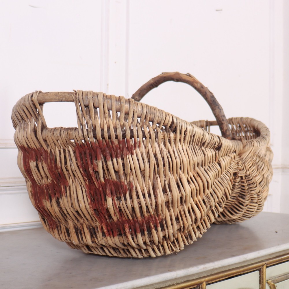 A large woven wicker twin harvest basket featuring a bentwood central handle and faded red painted marks on the natural exterior.