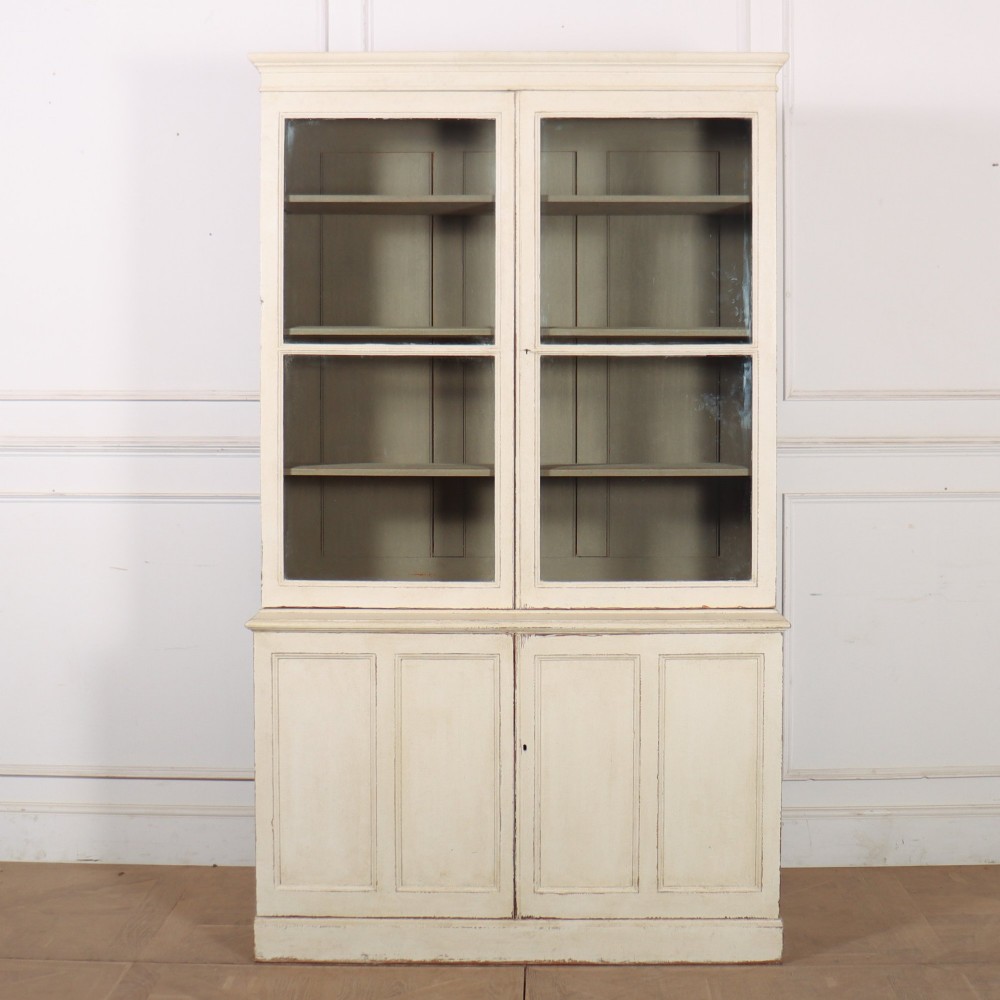 A tall 19th-century painted wooden bookcase featuring a cream exterior, two upper glazed doors revealing a grey interior with shelves, and a double-panelled lower cupboard base.