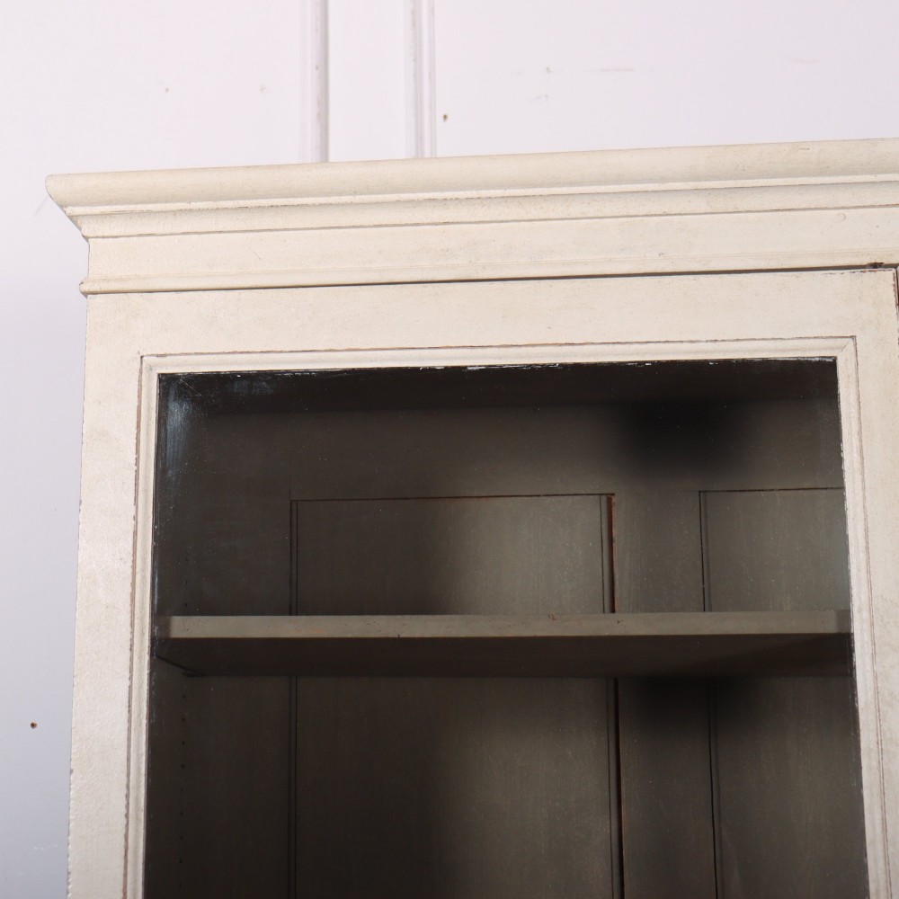 A tall 19th-century painted wooden bookcase featuring a cream exterior, two upper glazed doors revealing a grey interior with shelves, and a double-panelled lower cupboard base. View II