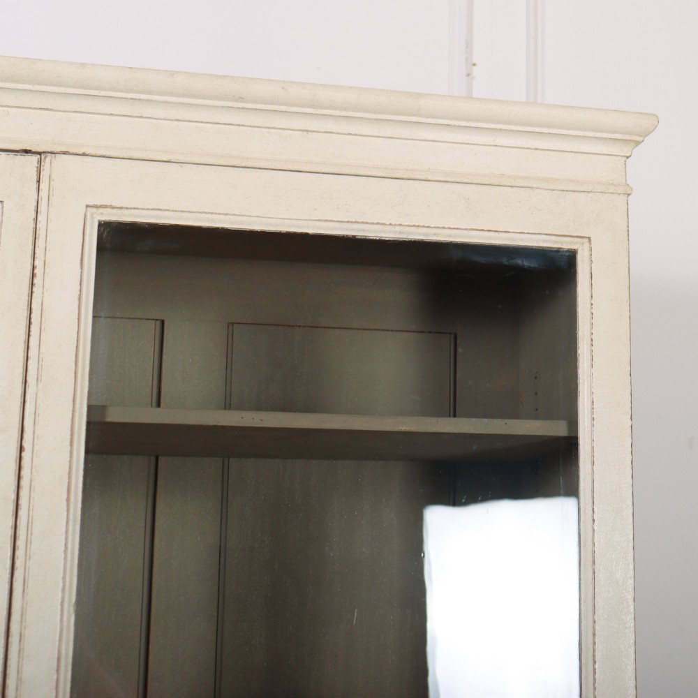A tall 19th-century painted wooden bookcase featuring a cream exterior, two upper glazed doors revealing a grey interior with shelves, and a double-panelled lower cupboard base. View III