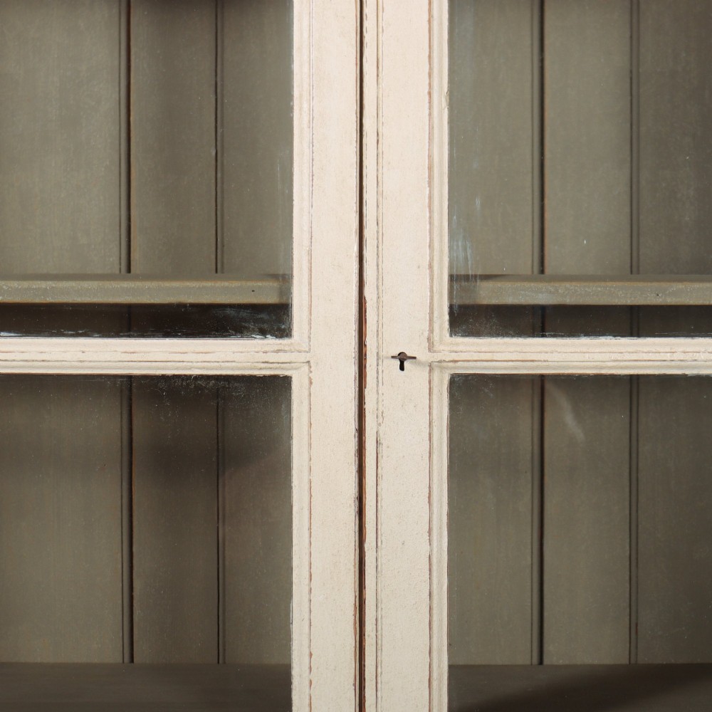 A tall 19th-century painted wooden bookcase featuring a cream exterior, two upper glazed doors revealing a grey interior with shelves, and a double-panelled lower cupboard base. View IV