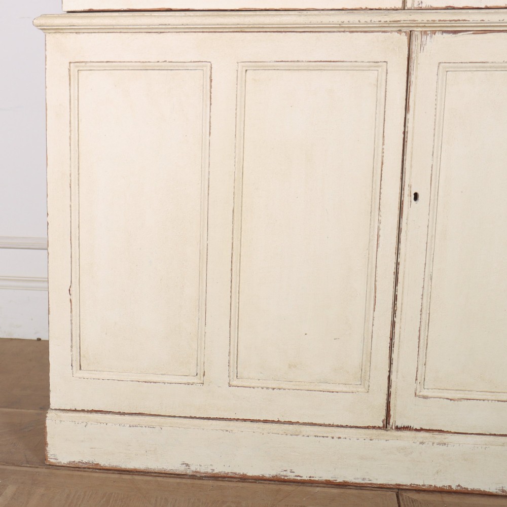 A tall 19th-century painted wooden bookcase featuring a cream exterior, two upper glazed doors revealing a grey interior with shelves, and a double-panelled lower cupboard base. View V