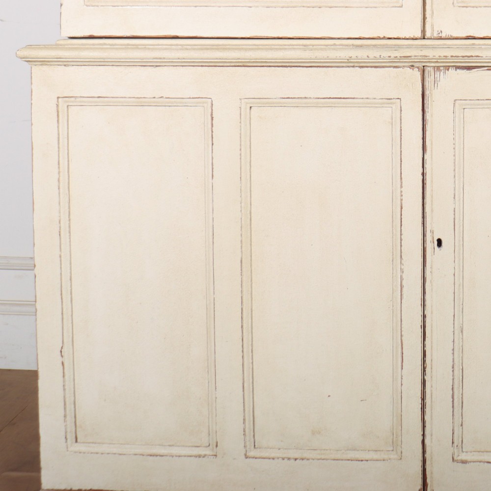 A tall 19th-century painted wooden bookcase featuring a cream exterior, two upper glazed doors revealing a grey interior with shelves, and a double-panelled lower cupboard base. View VI