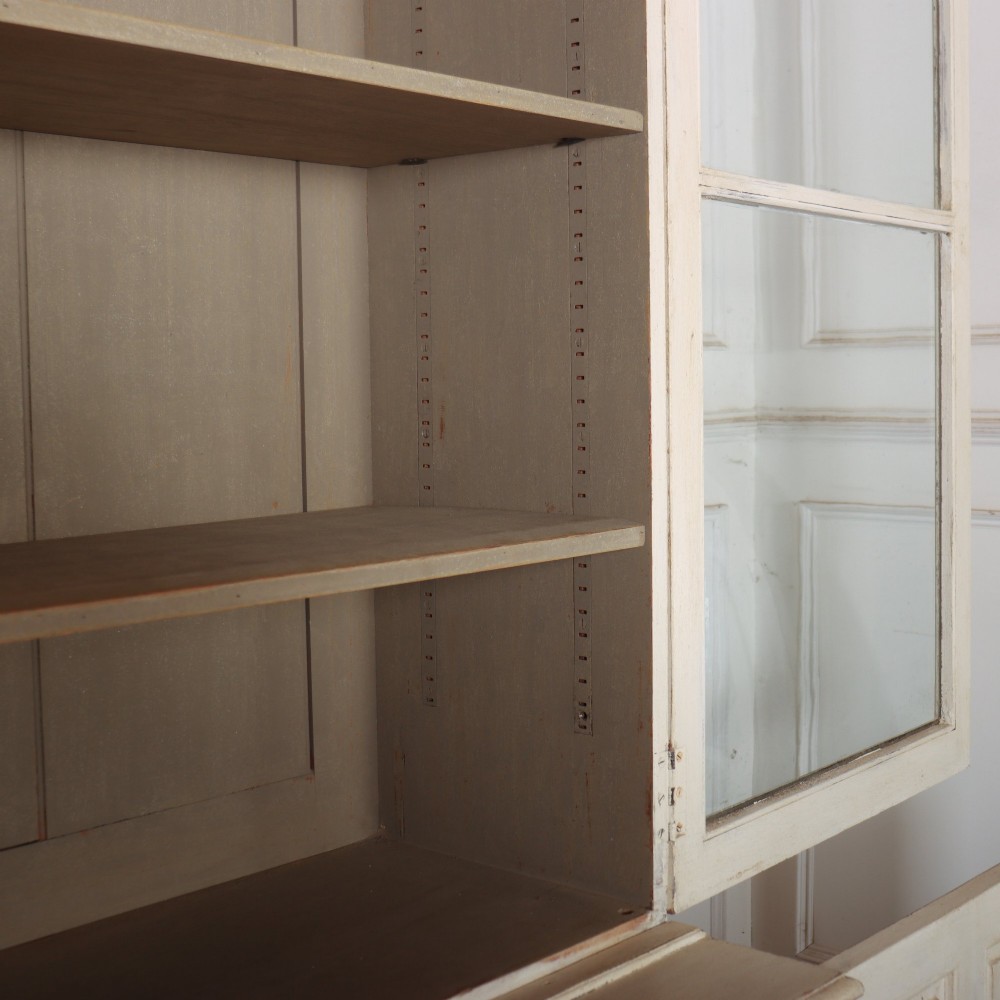 A tall 19th-century painted wooden bookcase featuring a cream exterior, two upper glazed doors revealing a grey interior with shelves, and a double-panelled lower cupboard base. View VIII