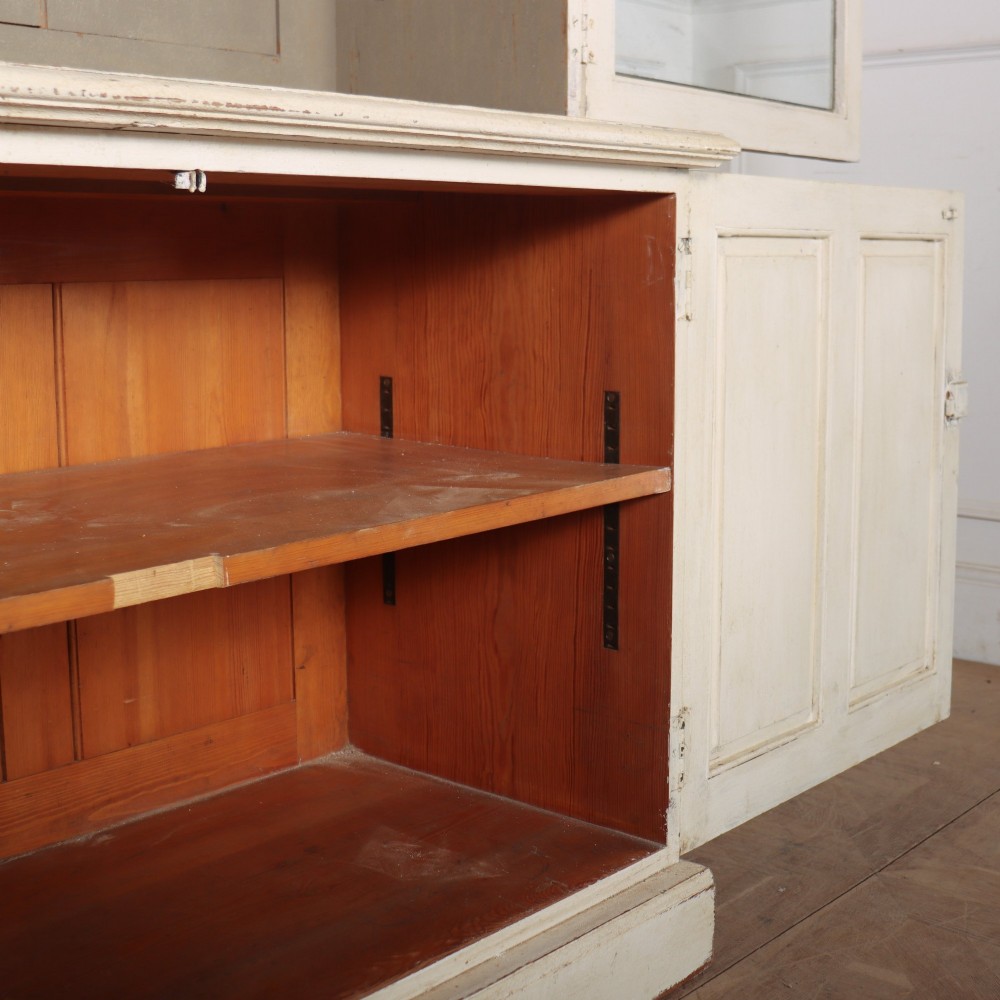 A tall 19th-century painted wooden bookcase featuring a cream exterior, two upper glazed doors revealing a grey interior with shelves, and a double-panelled lower cupboard base. View IX
