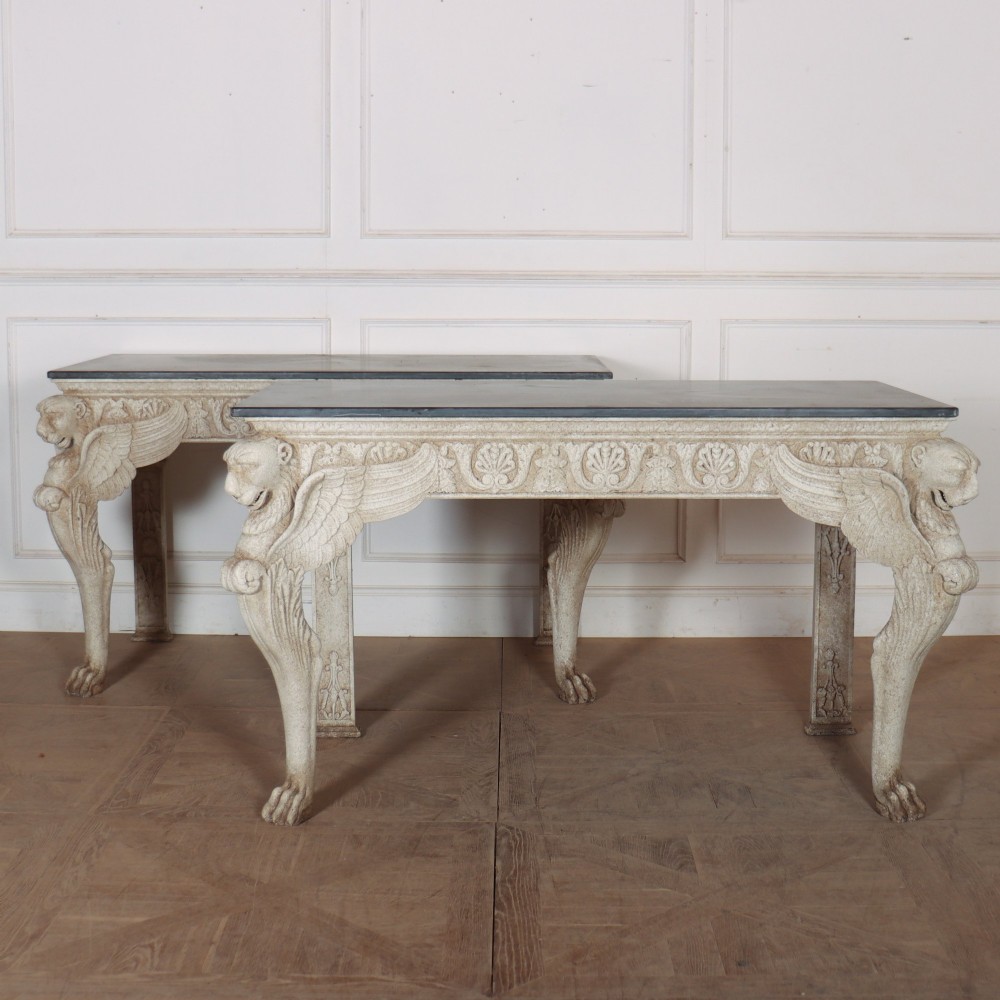 A pair of large off-white painted console tables featuring carved winged lion monopodium supports, anthemion friezes, and dark grey tops.