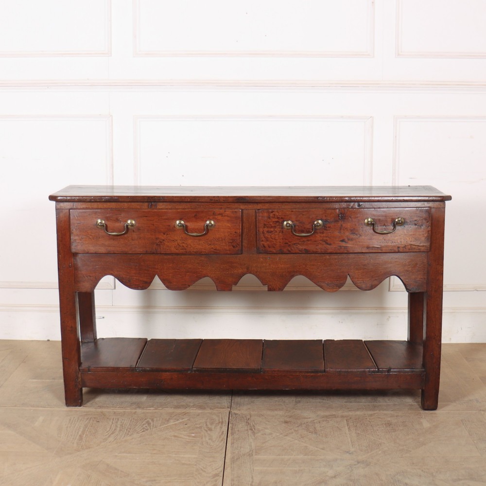 An antique dark oak Welsh dresser base featuring two broad drawers with brass handles, a decorative scalloped apron, and an open lower pot board shelf.