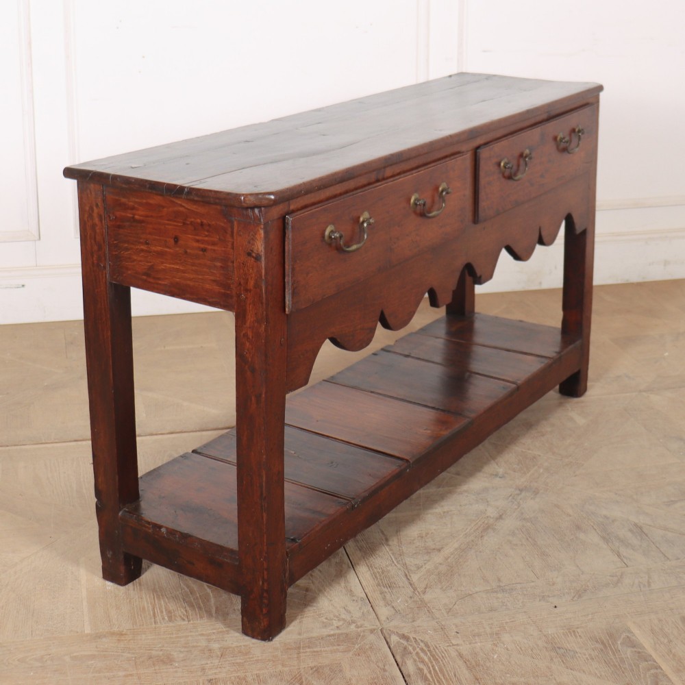 An antique dark oak Welsh dresser base featuring two broad drawers with brass handles, a decorative scalloped apron, and an open lower pot board shelf. View VII