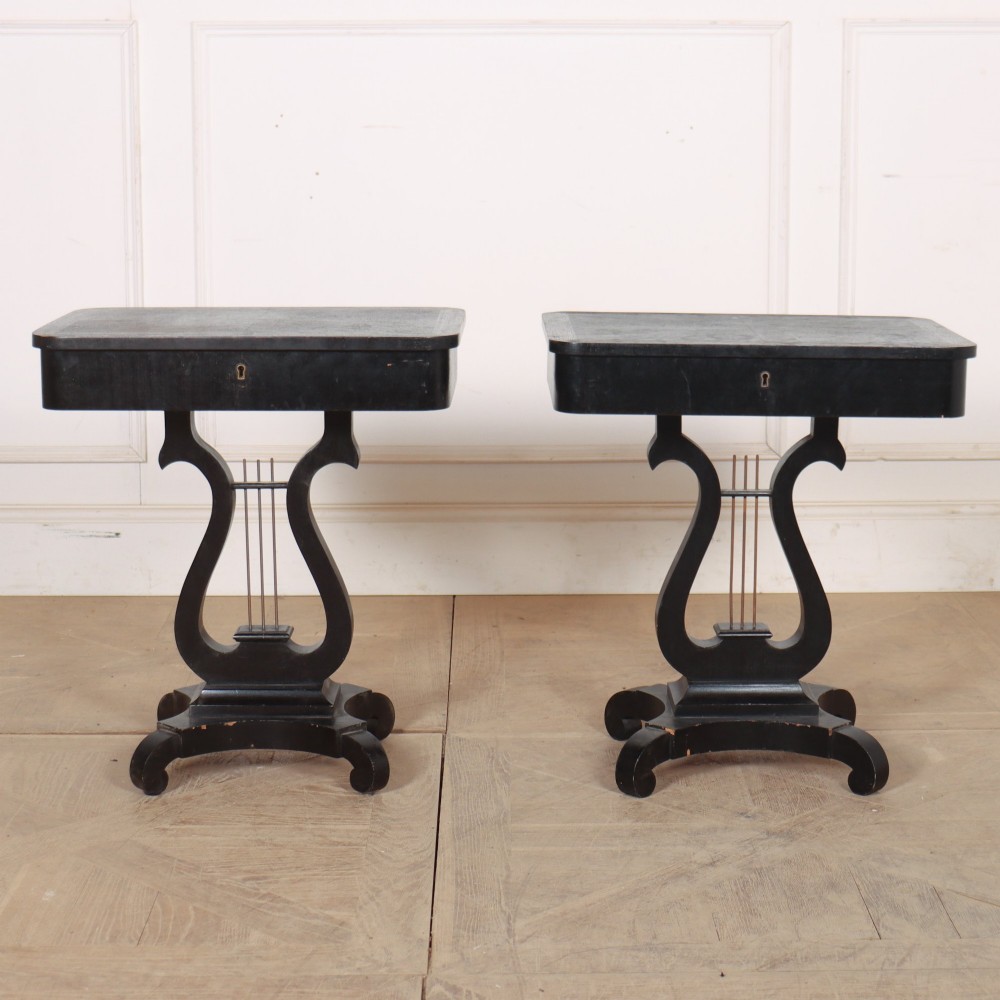 A pair of black ebonised side tables featuring single drawers with brass keyholes and lyre-shaped pedestal bases resting on scroll feet.