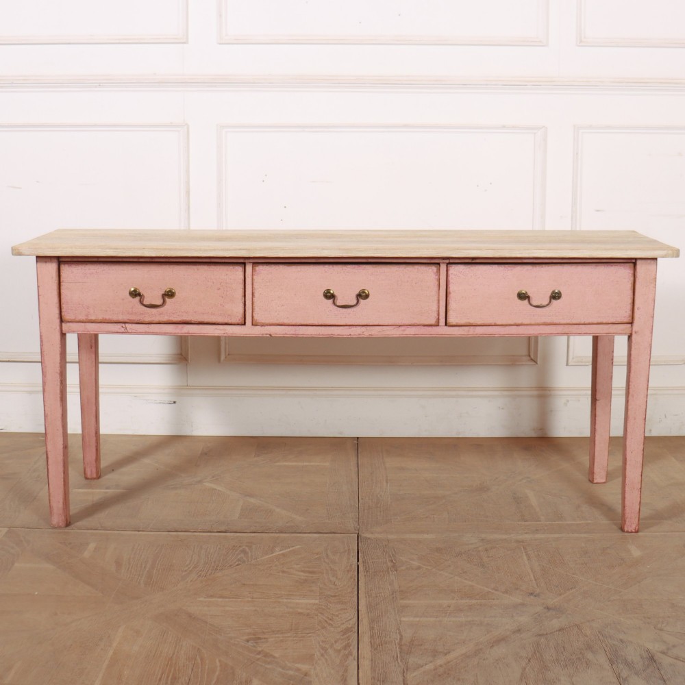 An antique English wooden server featuring a scrubbed natural timber top, a distressed pink painted frame, three drawers with brass drop handles, and four straight legs.