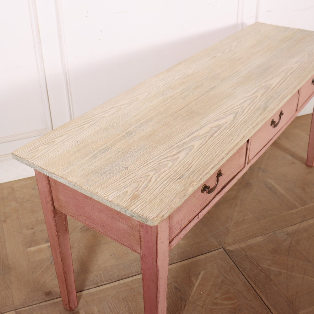 An antique English wooden server featuring a scrubbed natural timber top, a distressed pink painted frame, three drawers with brass drop handles, and four straight legs. View X