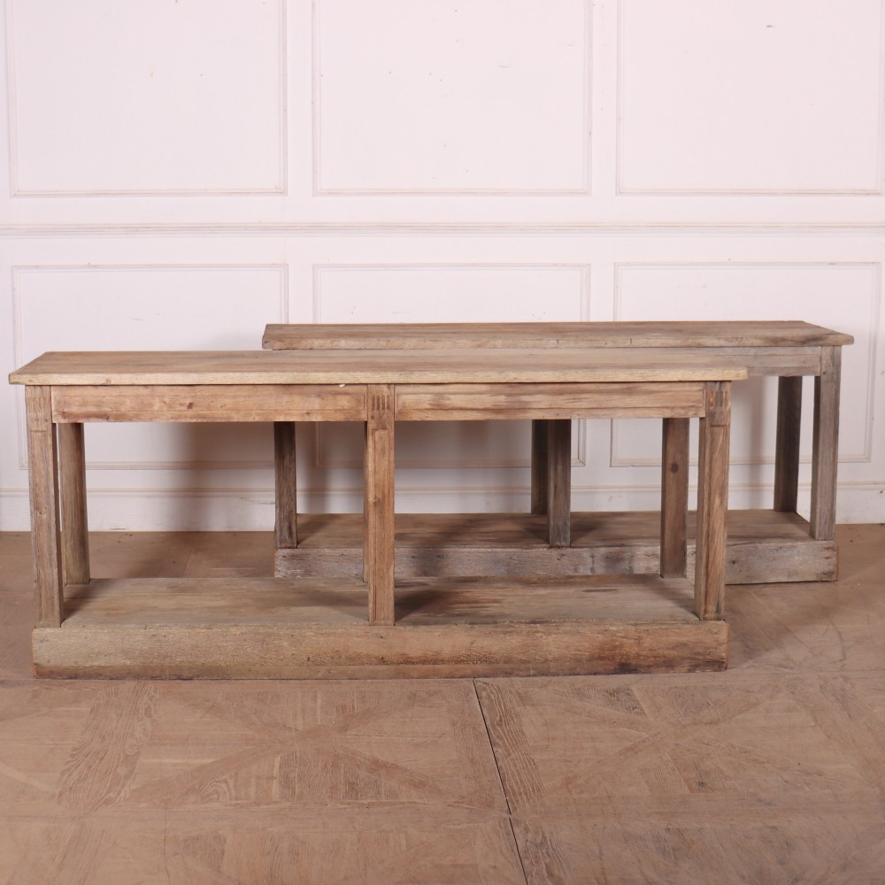 A pair of long rustic wooden French console tables with lower open shelves and weathered pale timber finish.