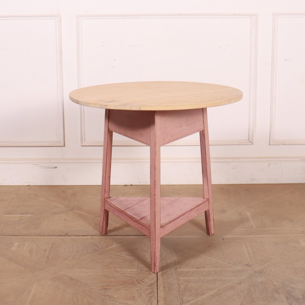 A nineteenth-century cricket table with a round, light natural wood top resting on a three-legged pink painted frame with a triangular lower shelf.