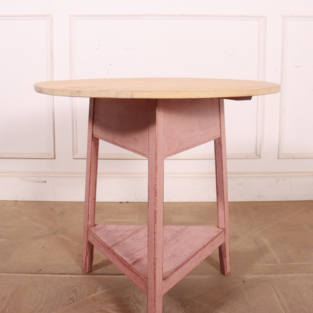 A nineteenth-century cricket table with a round, light natural wood top resting on a three-legged pink painted frame with a triangular lower shelf. View III