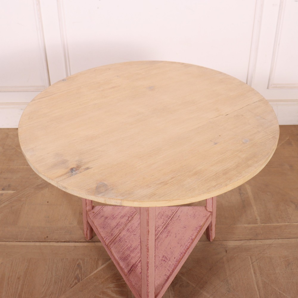 A nineteenth-century cricket table with a round, light natural wood top resting on a three-legged pink painted frame with a triangular lower shelf. View IV
