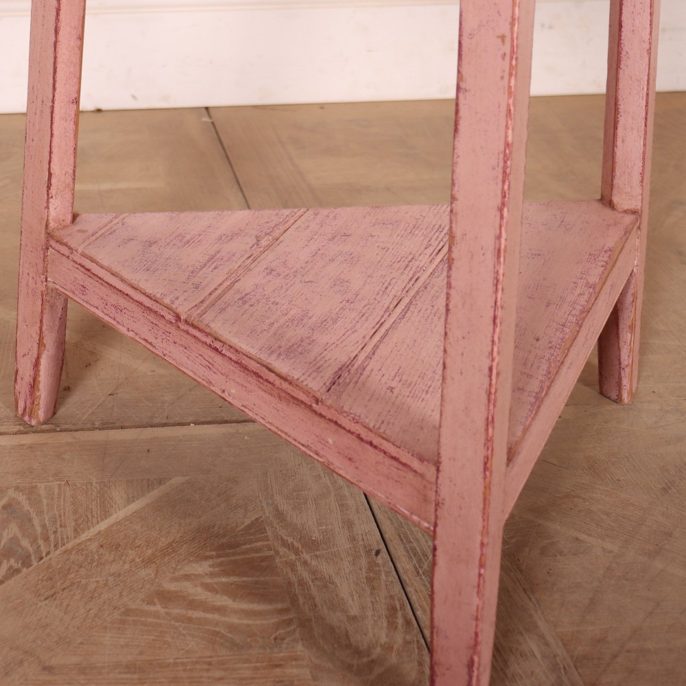 A nineteenth-century cricket table with a round, light natural wood top resting on a three-legged pink painted frame with a triangular lower shelf. View V