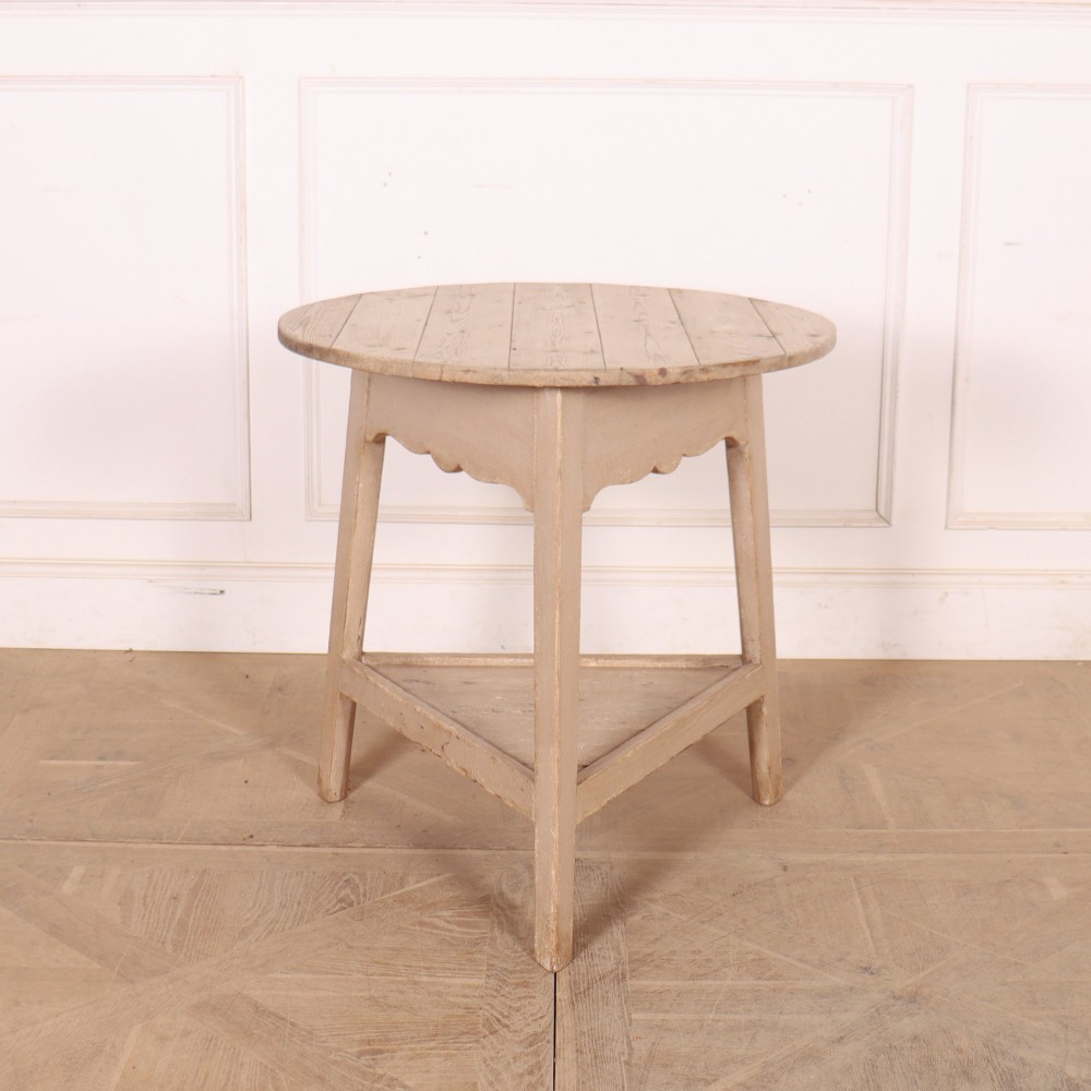 A nineteenth-century round wooden cricket table with three tapered legs, a lower triangular shelf, a scalloped apron, and a pale grey painted finish.