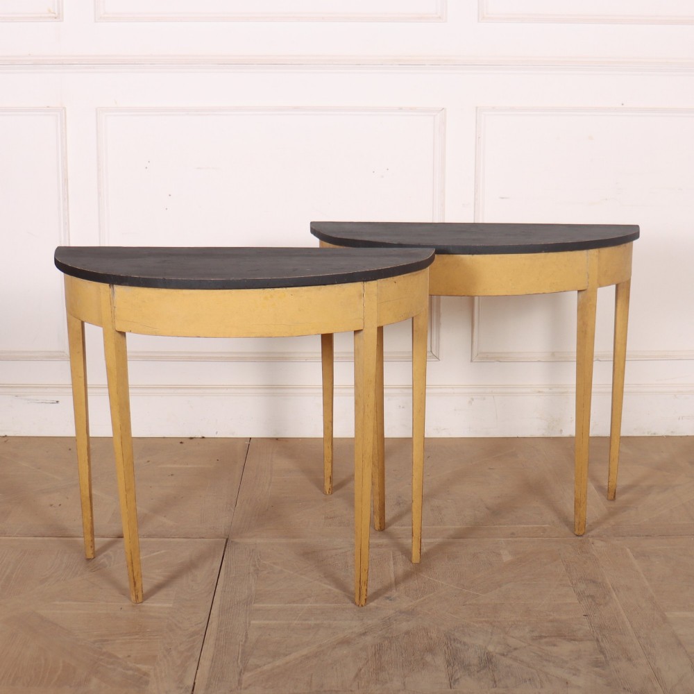 A pair of small painted demi-lune console tables with black tops, ochre yellow bases, and slender square tapered legs.