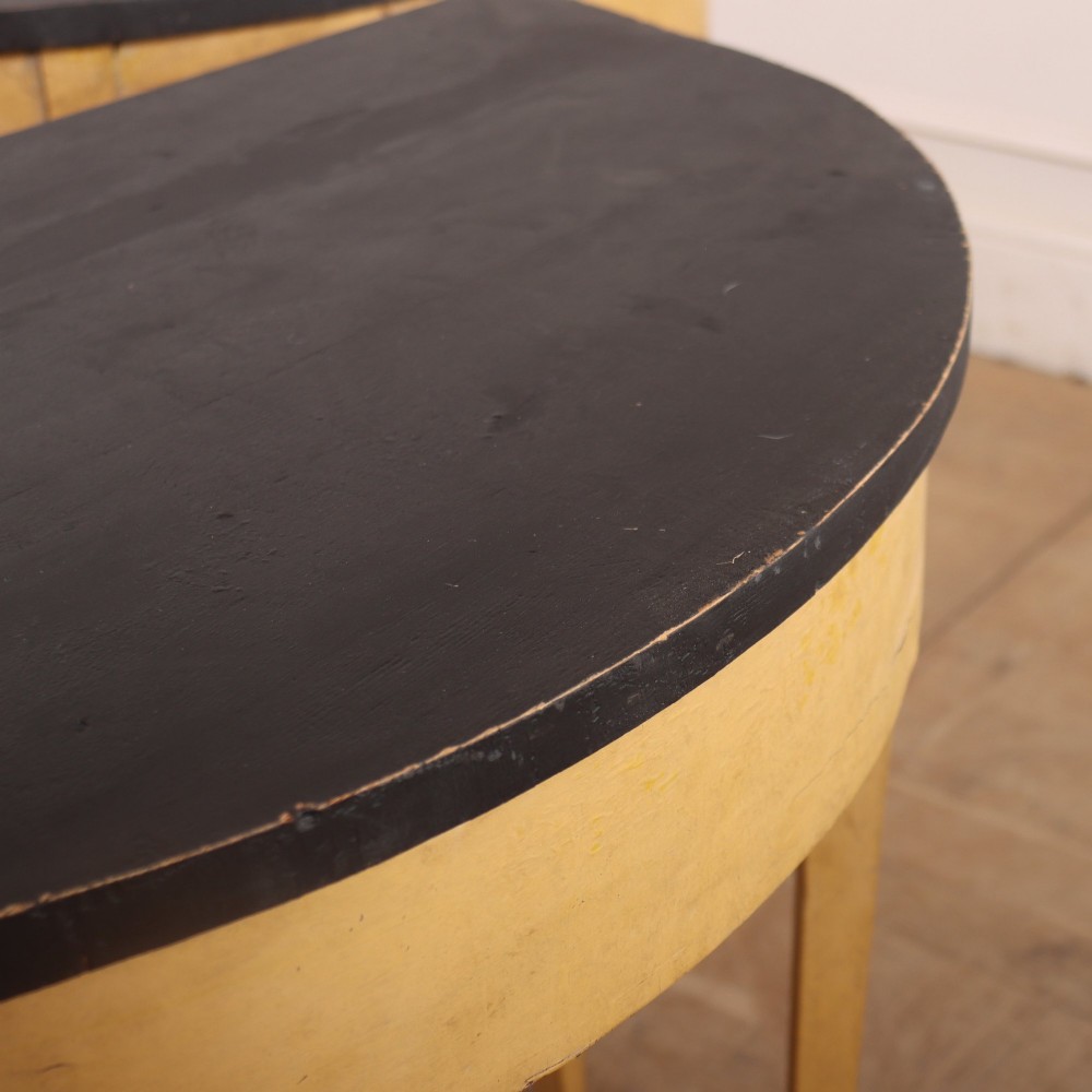 A pair of small painted demi-lune console tables with black tops, ochre yellow bases, and slender square tapered legs. View VI