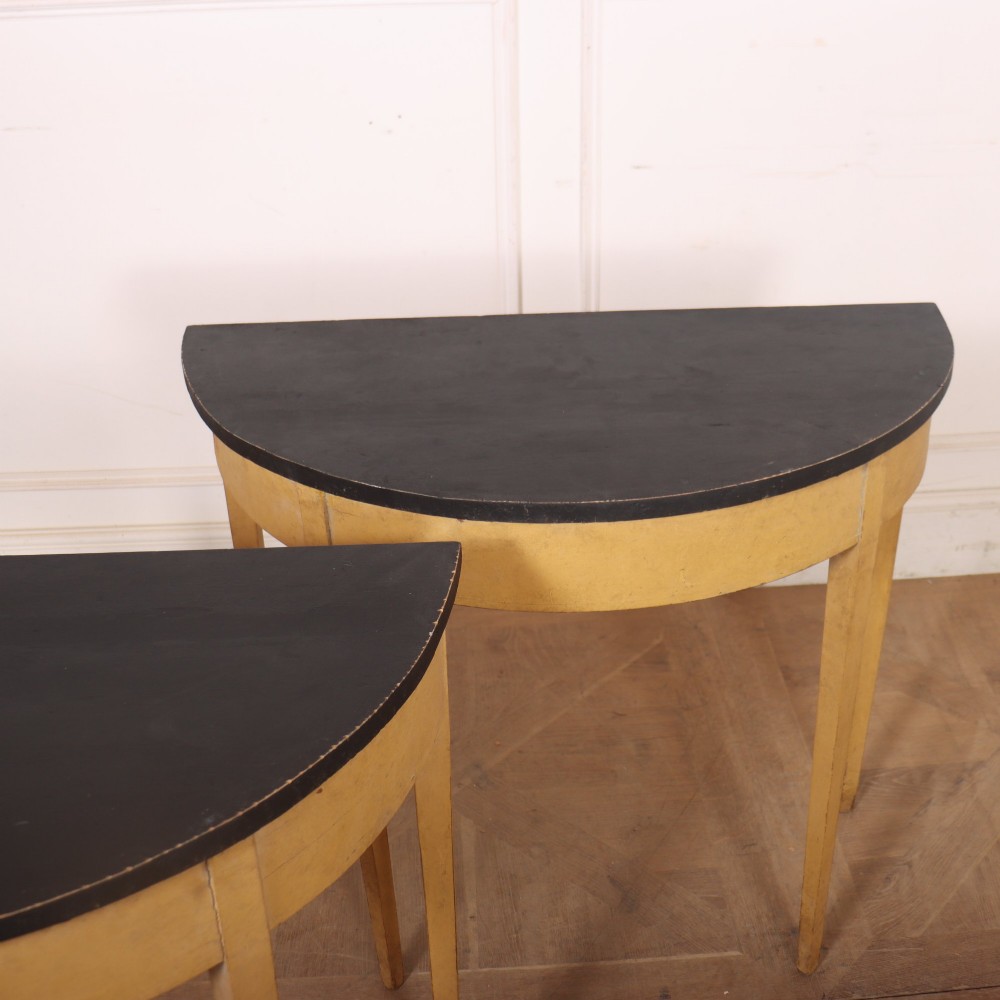 A pair of small painted demi-lune console tables with black tops, ochre yellow bases, and slender square tapered legs. View VII
