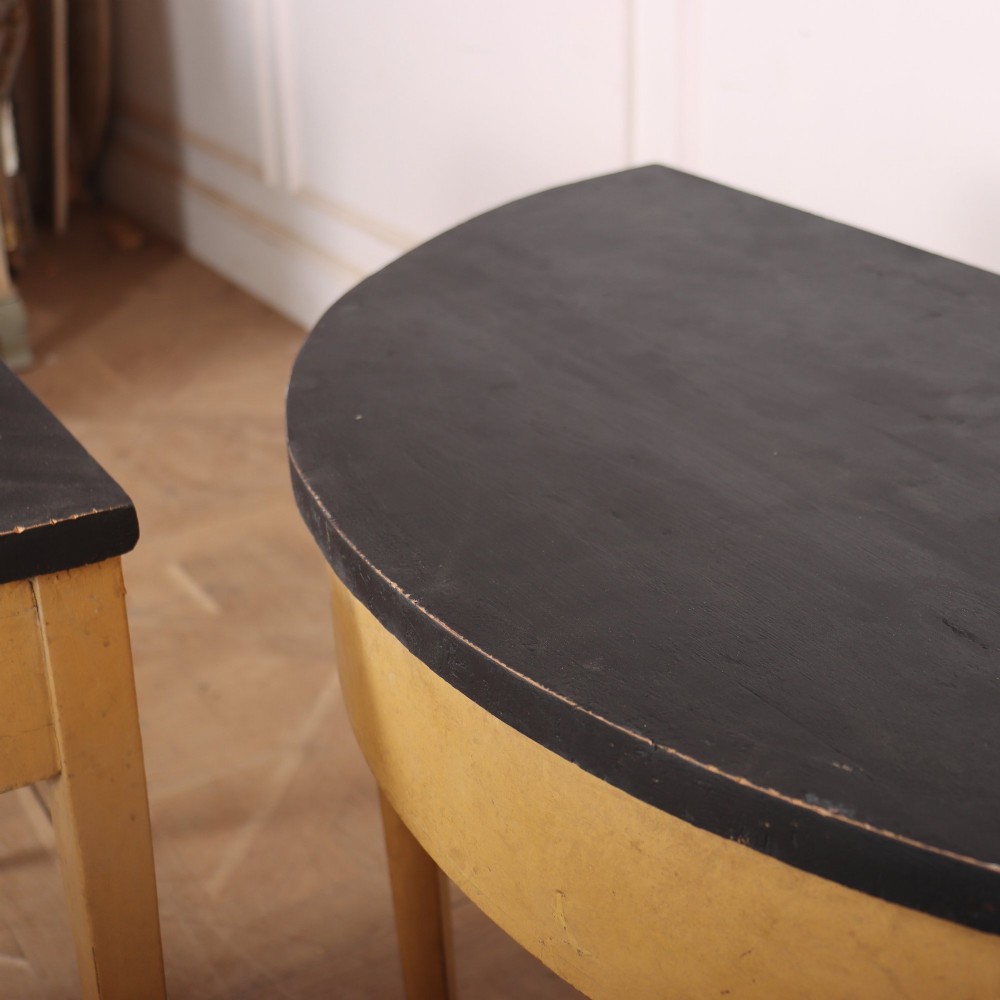 A pair of small painted demi-lune console tables with black tops, ochre yellow bases, and slender square tapered legs. View VIII
