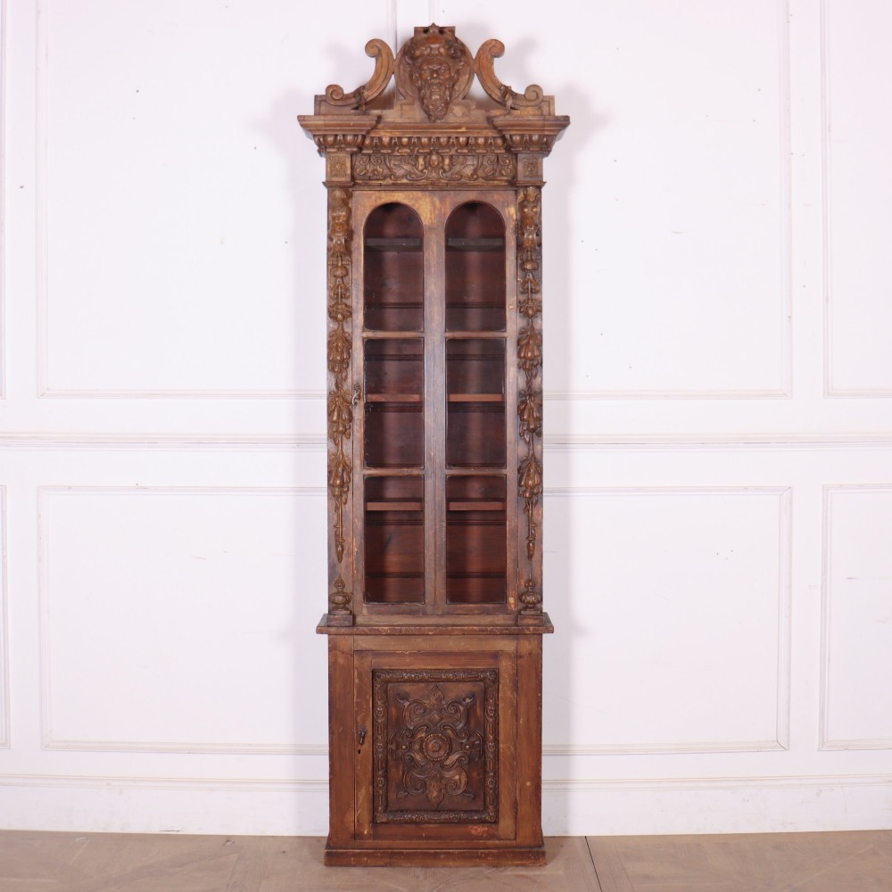 A tall, narrow carved pine chimney cupboard featuring a decorative carved crest, two glazed upper doors, and a carved lower door panel.
