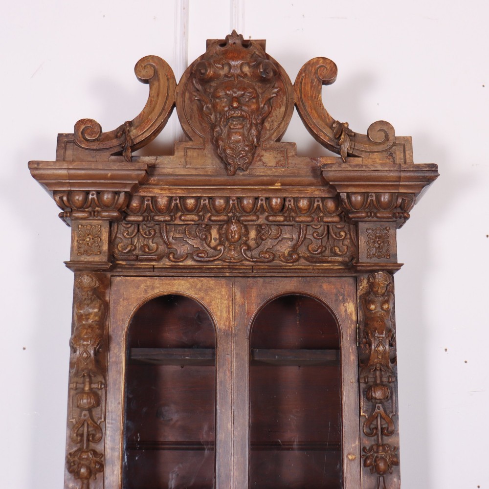 A tall, narrow carved pine chimney cupboard featuring a decorative carved crest, two glazed upper doors, and a carved lower door panel. View II