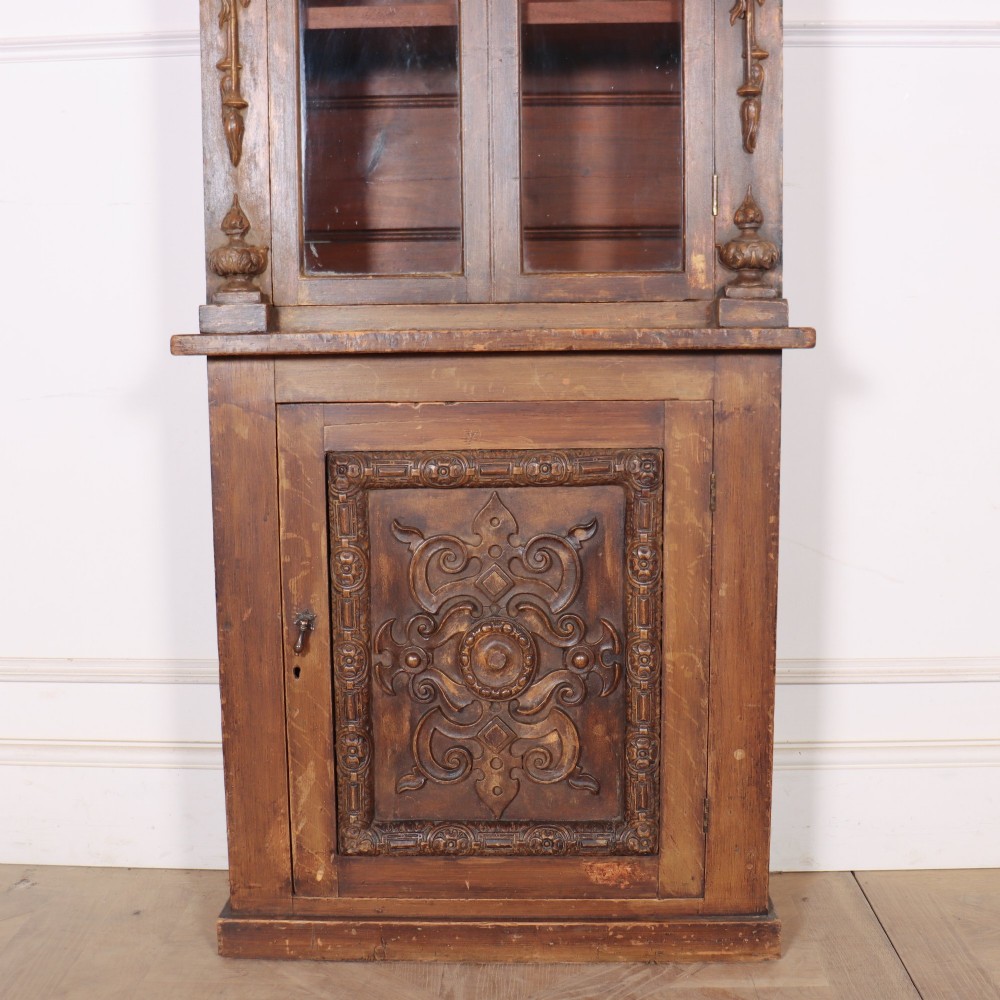 A tall, narrow carved pine chimney cupboard featuring a decorative carved crest, two glazed upper doors, and a carved lower door panel. View III