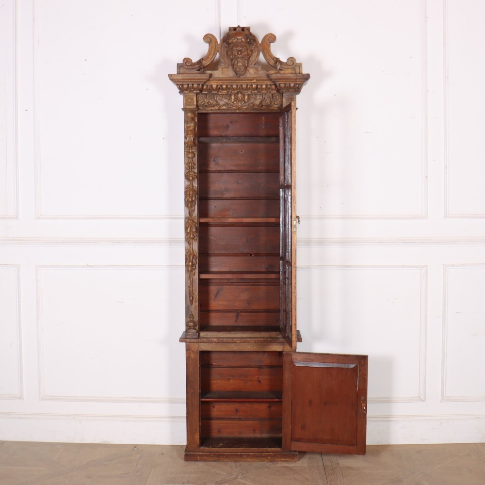 A tall, narrow carved pine chimney cupboard featuring a decorative carved crest, two glazed upper doors, and a carved lower door panel. View VI