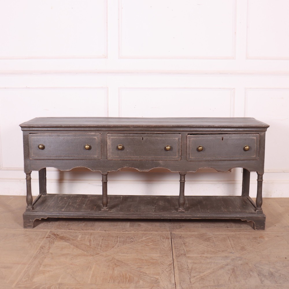 An English painted potboard dresser base in slate grey featuring three frieze drawers with brass handles and a low open shelf supported by turned legs.