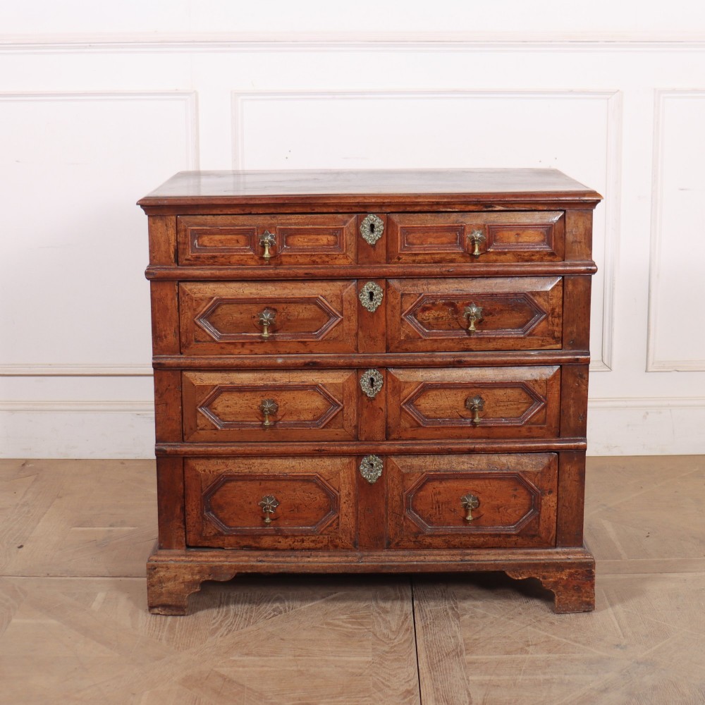 An eighteenth-century oak chest of drawers featuring geometric panelled drawer fronts, brass drop handles, brass escutcheons, and bracket feet.