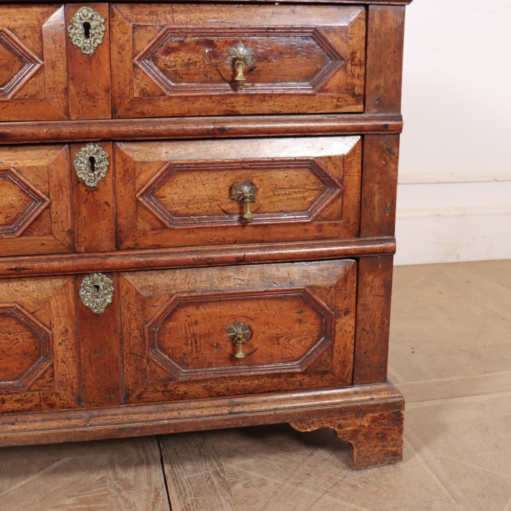 An eighteenth-century oak chest of drawers featuring geometric panelled drawer fronts, brass drop handles, brass escutcheons, and bracket feet. View V