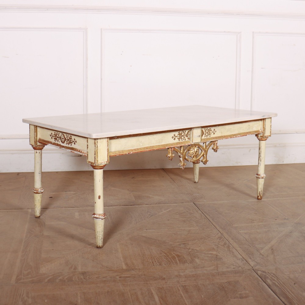 An Italian Neoclassical low coffee table featuring a flat white marble top, a cream painted apron with carved gilded accents, and tapered legs.