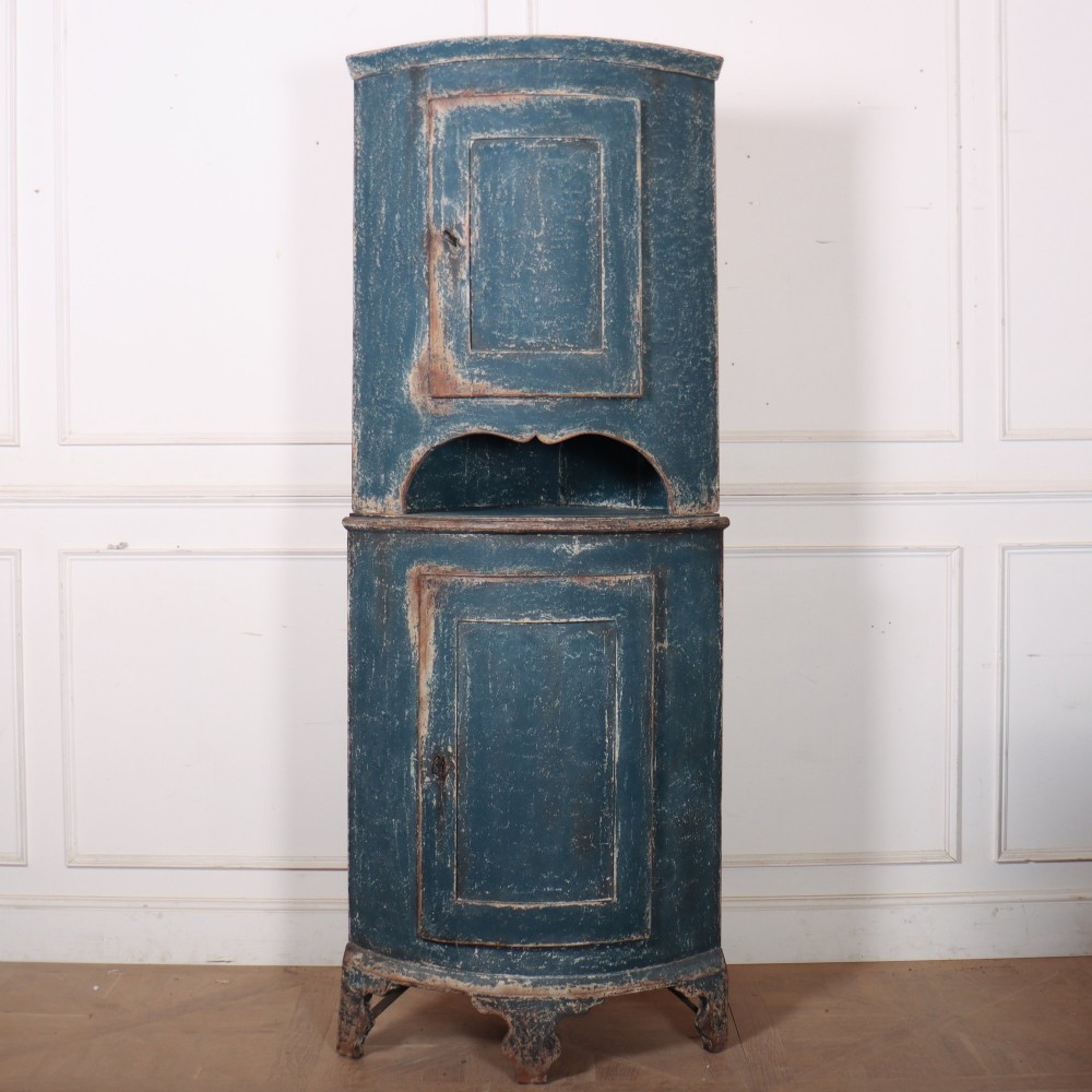A tall Swedish bowfronted corner cupboard with a distressed blue painted finish, featuring upper and lower panel doors with an open middle shelf.