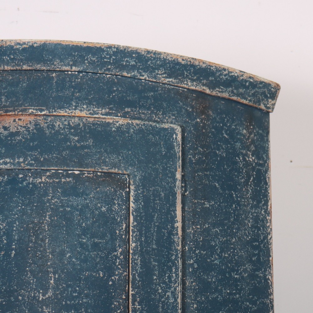 A tall Swedish bowfronted corner cupboard with a distressed blue painted finish, featuring upper and lower panel doors with an open middle shelf. View III