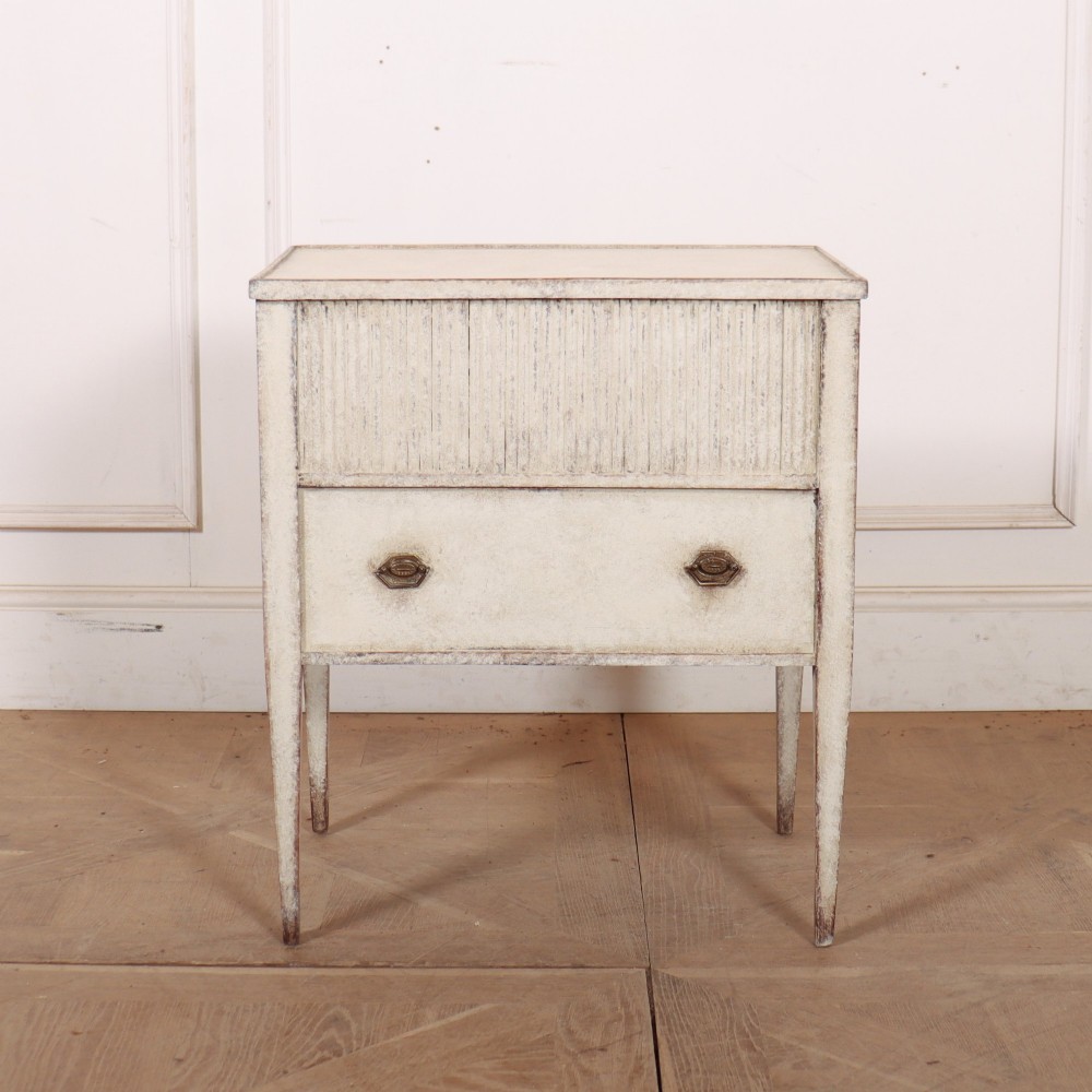 An antique cream painted Dutch bedside cabinet featuring a fluted upper section, a lower drawer with brass handles, and square tapered legs.