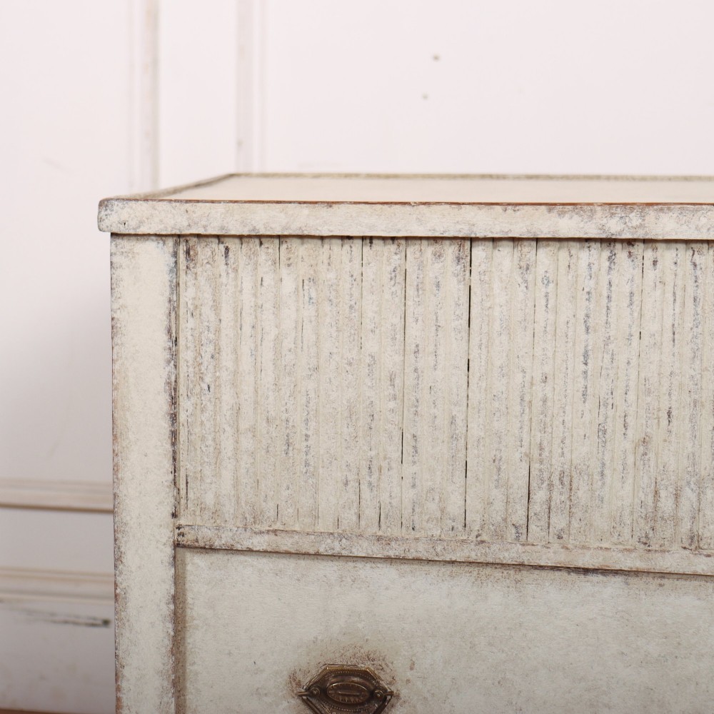 An antique cream painted Dutch bedside cabinet featuring a fluted upper section, a lower drawer with brass handles, and square tapered legs. View II