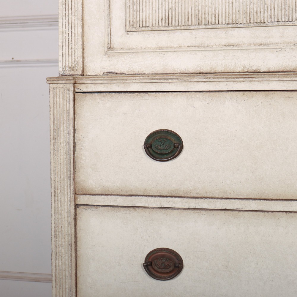 A tall off-white painted Scandinavian style linen cupboard featuring a two-door upper section above three drawers with circular brass handles. View V