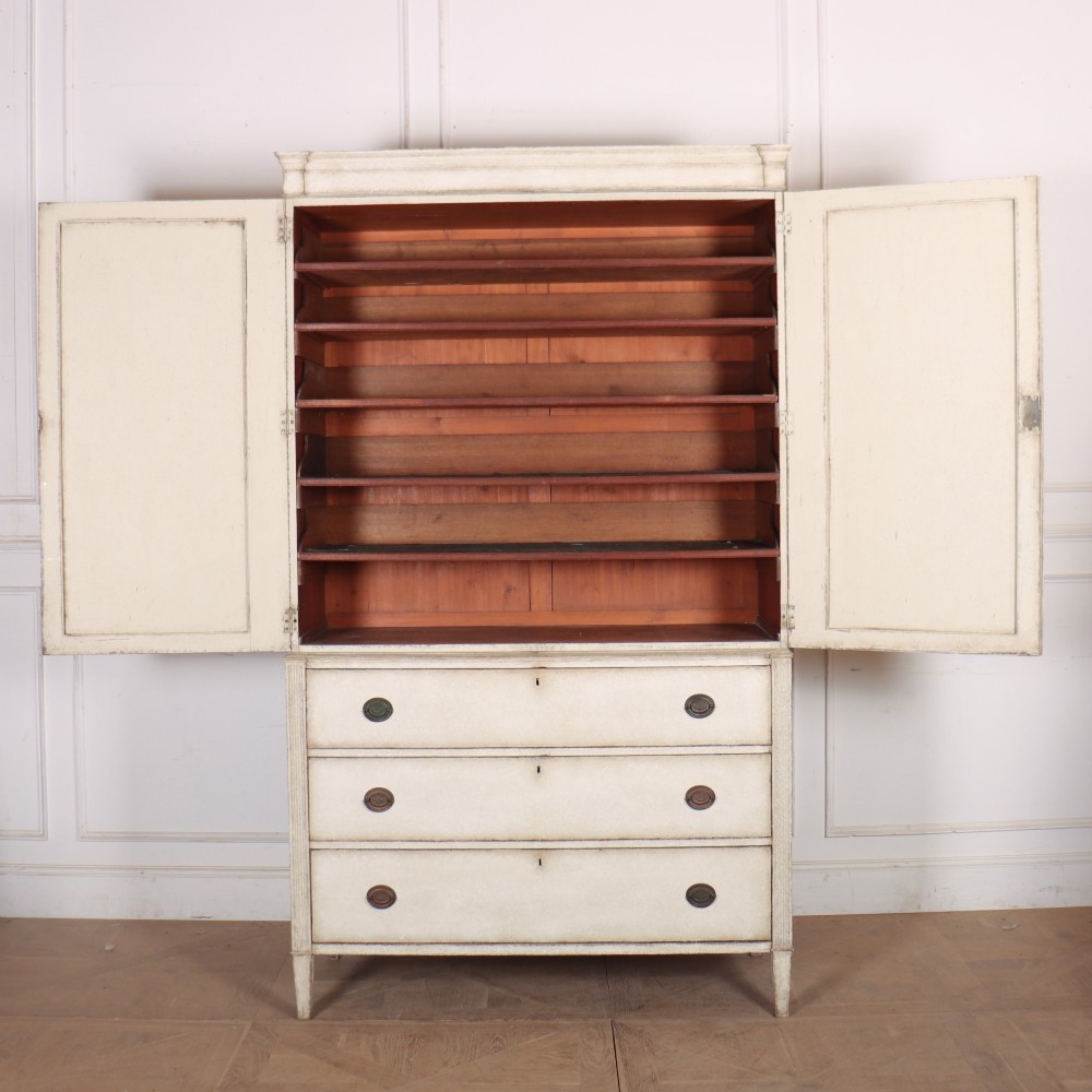 A tall off-white painted Scandinavian style linen cupboard featuring a two-door upper section above three drawers with circular brass handles. View X
