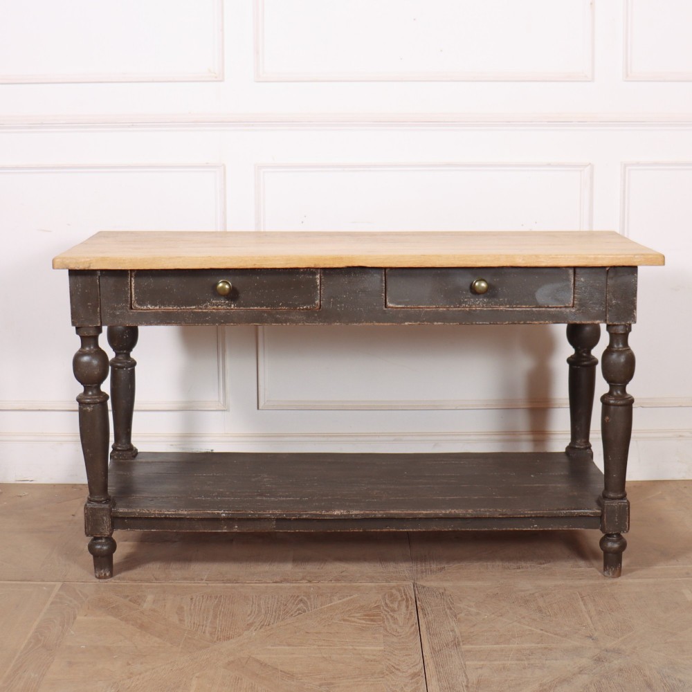 A French drapers table featuring a natural wood top, two shallow drawers with brass knobs, a dark grey painted base, turned legs, and a lower pot board shelf.