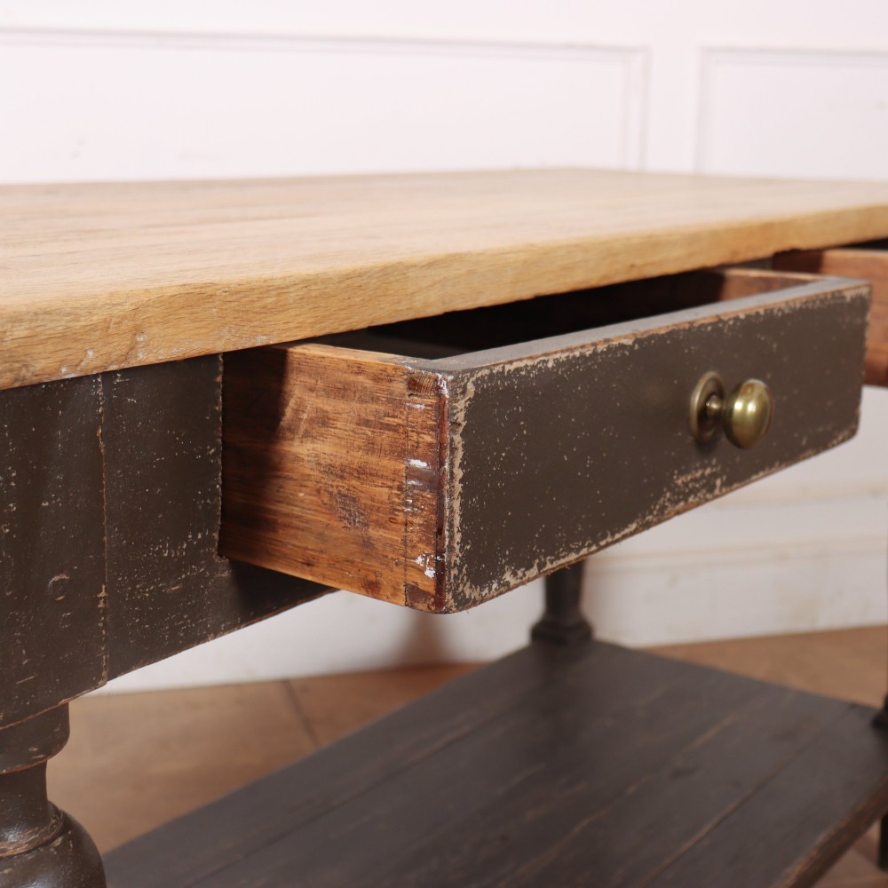 A French drapers table featuring a natural wood top, two shallow drawers with brass knobs, a dark grey painted base, turned legs, and a lower pot board shelf. View X