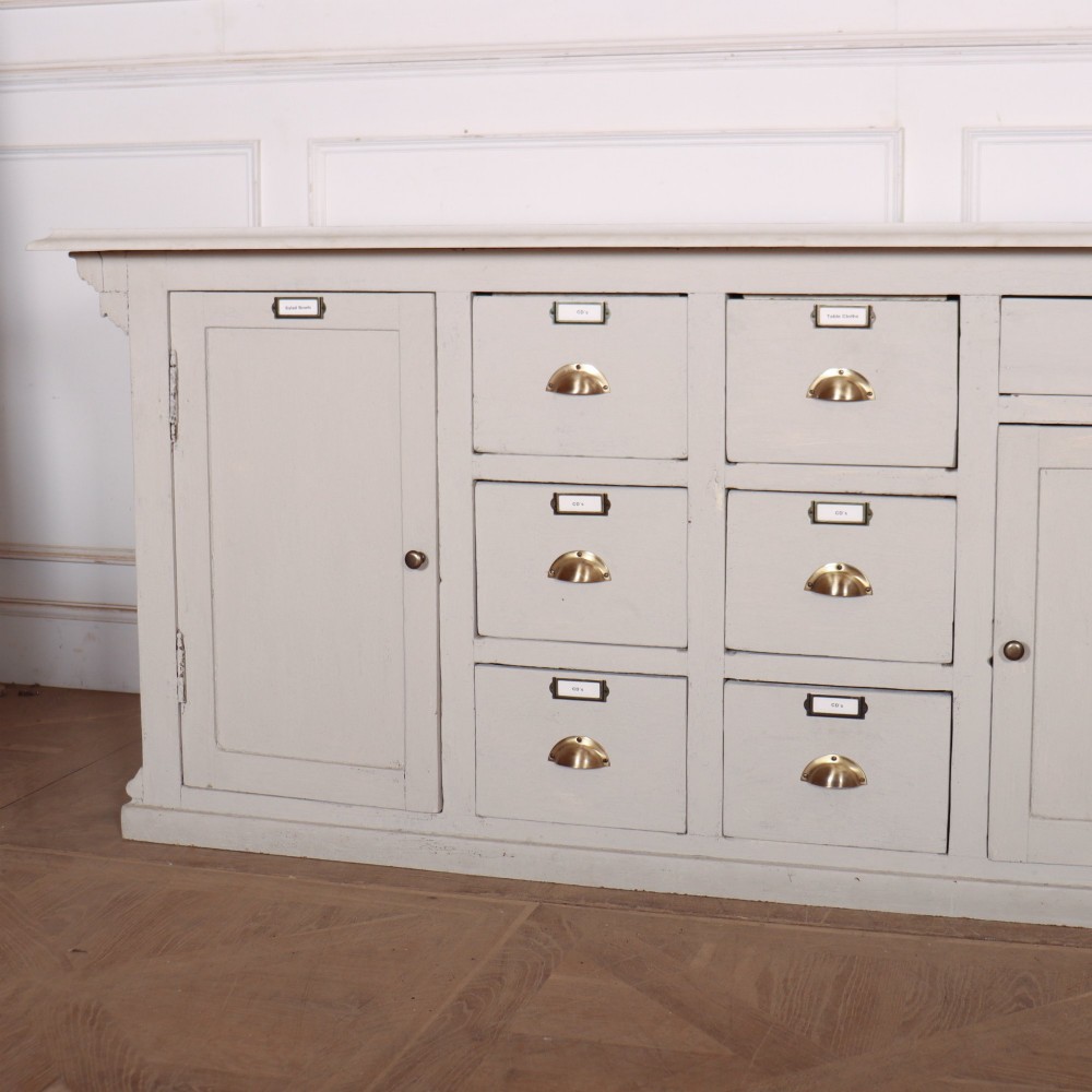 A long off-white painted timber shop counter featuring fluted Corinthian pilasters and recessed panel moulding along the front. View XIII