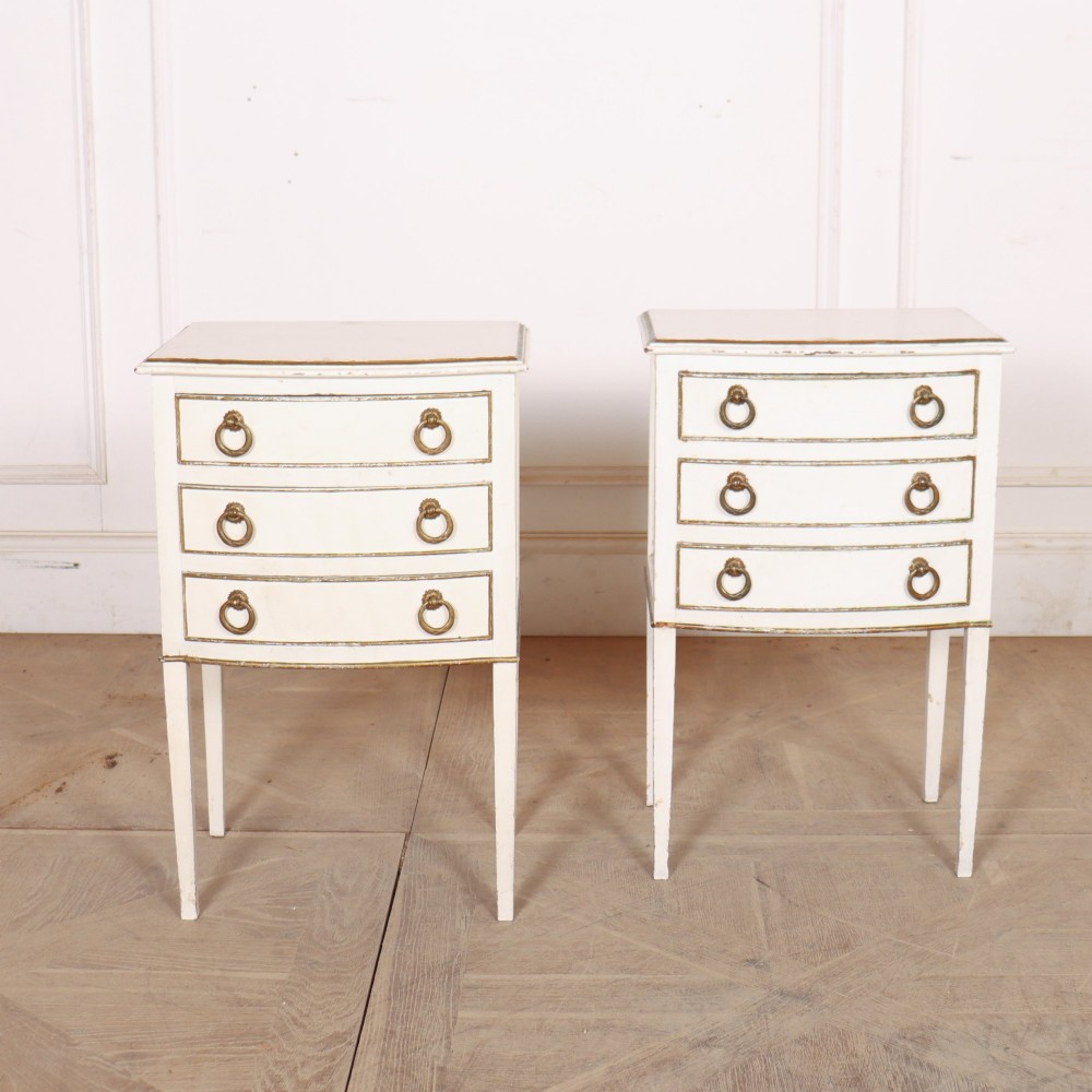 A pair of off-white painted French bedside chests featuring three curved drawers, gilded moulding details, brass ring handles, and tall square tapered legs.